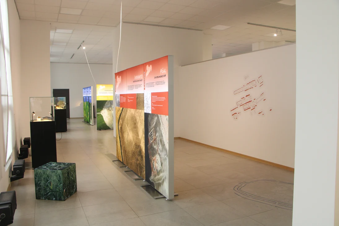 Leuchtwände zeigen Luftbilder und Grafiken im Themenbereich "Siedlungen". Im Hintergrund ist eine große Grafik auf der weißen Wand zu sehen. Vor den Fenstern stehen Vitrinen.