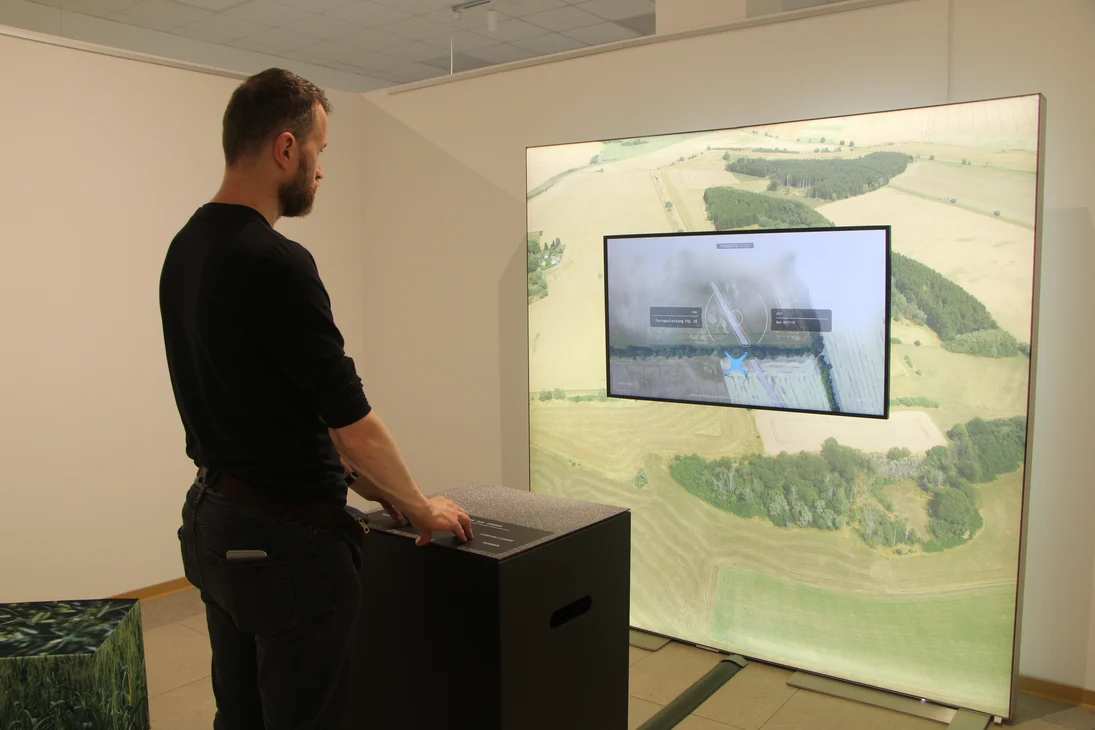 Ein Mann steht vor einer Spielekonsole und lenkt eine virtuelle Drohne über großes Luftbild vor ihm. Mit dem Drohnenspiel für die Besucherinnen und Besucher. Hier kann man spielerisch Spuren aus der Luft entdecken.