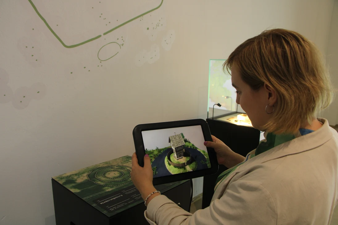 An einer Konsole hält eine Frau ein Tablet, auf dem ein dreidimensionales Bild einer Turmhügelburg erscheint, sobald sie damit auf das Foto auf der Konsole zeigt. Die Burg besteht aus einem Holzturm auf einem künstlichen Hügel umgeben von einem Graben.