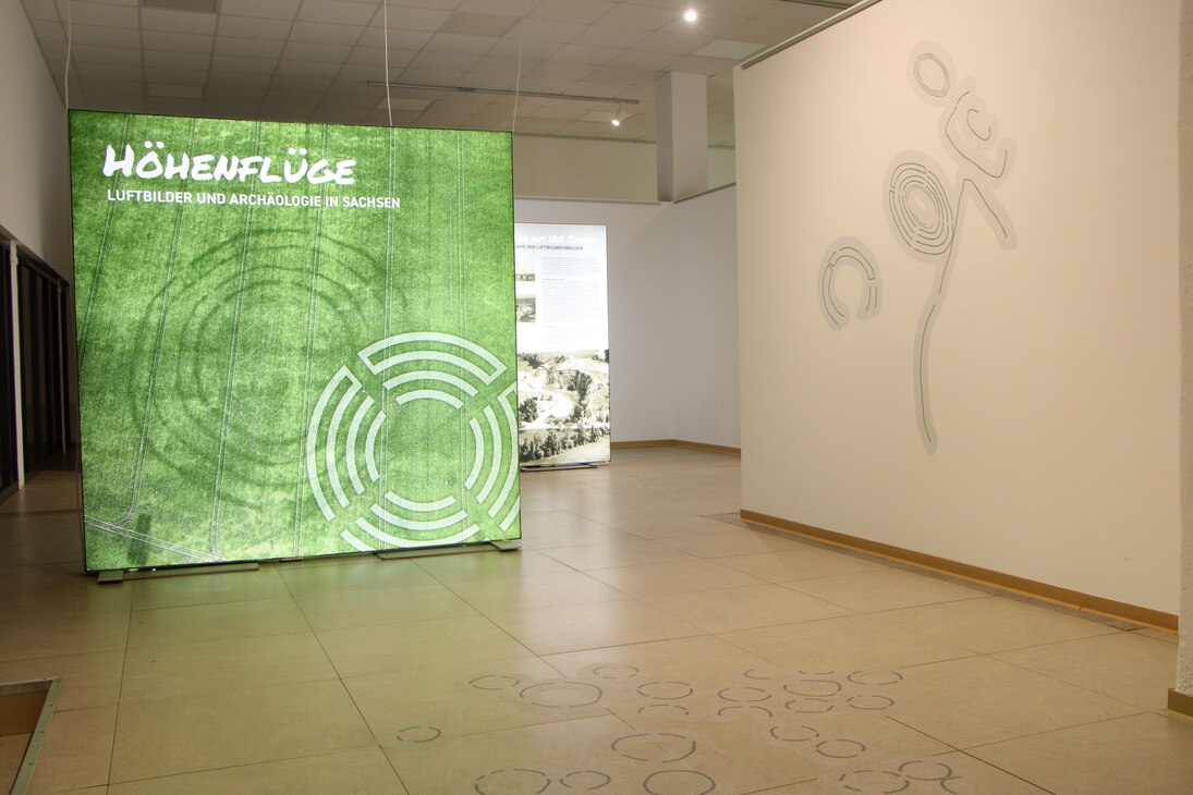 Titelaufsteller der Ausstellung "Höhenflüge. Luftbilder und Archäologie in Sachsen" mit einem Bild der Kreisgrabenanlage von Kyhna