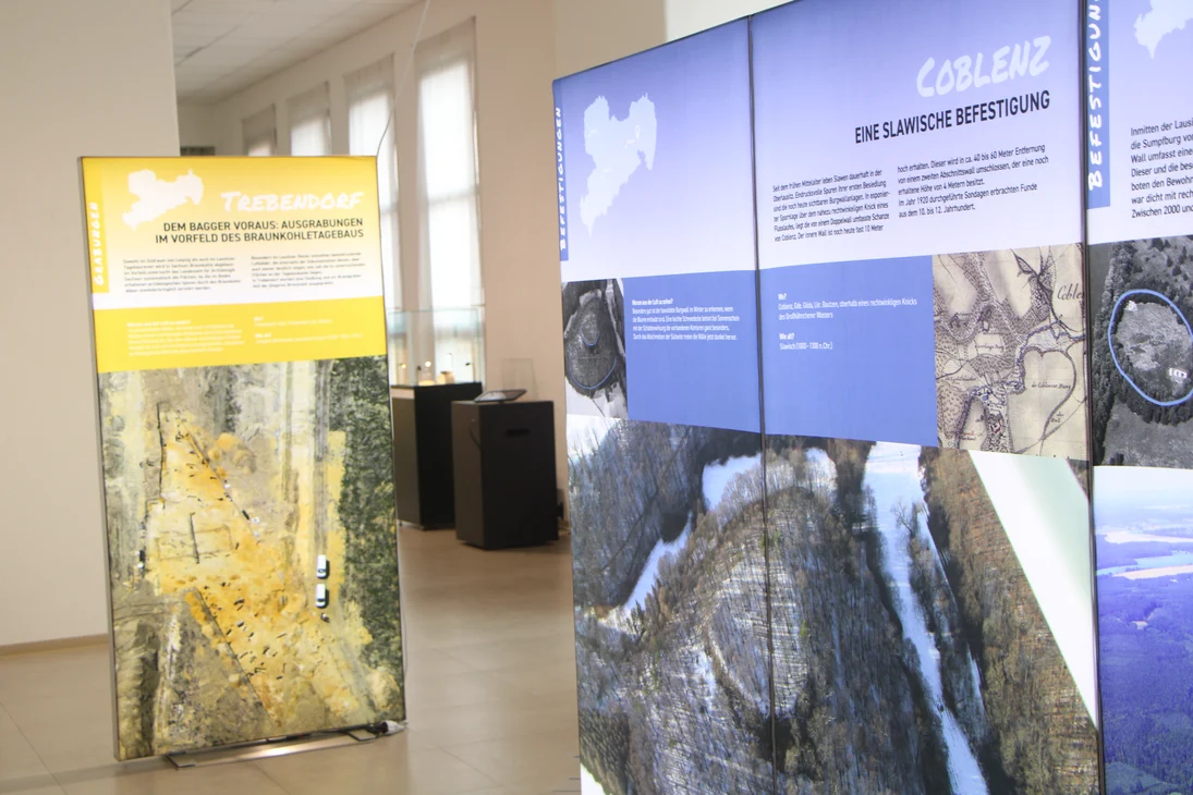 Dieser Blick in die Ausstellung zeigt im Vordergrund eine Große Blaue Leuchtwand mit einem winterlichen Luftbild der slawischen Befestigung von Coblenz. Die kleinere gelbe Wand im Hintergrund zeigt ein Bild vom Braunkohletagebau Nochten bei Trebendorf.
