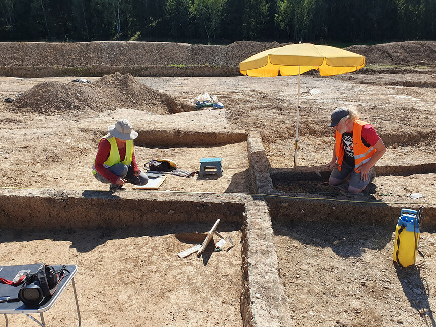 Zwei Archäologen sind auf der Grabung bei Rodau bei der Arbeit, einer unter einem gelben Sonnenschirm. 